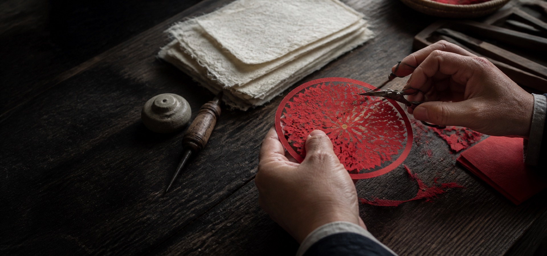 Hands carefully cutting a detailed red paper motif beside traditional craft tools.