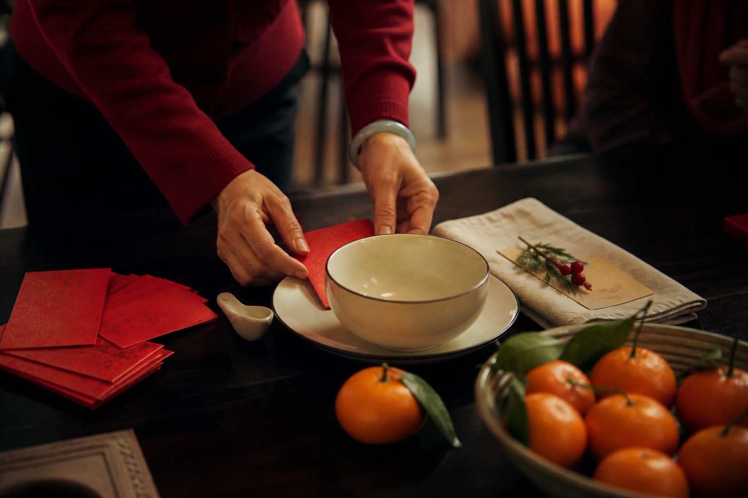 Hands arranging red envelopes, citrus fruit, and a family place setting for Lunar New Year.