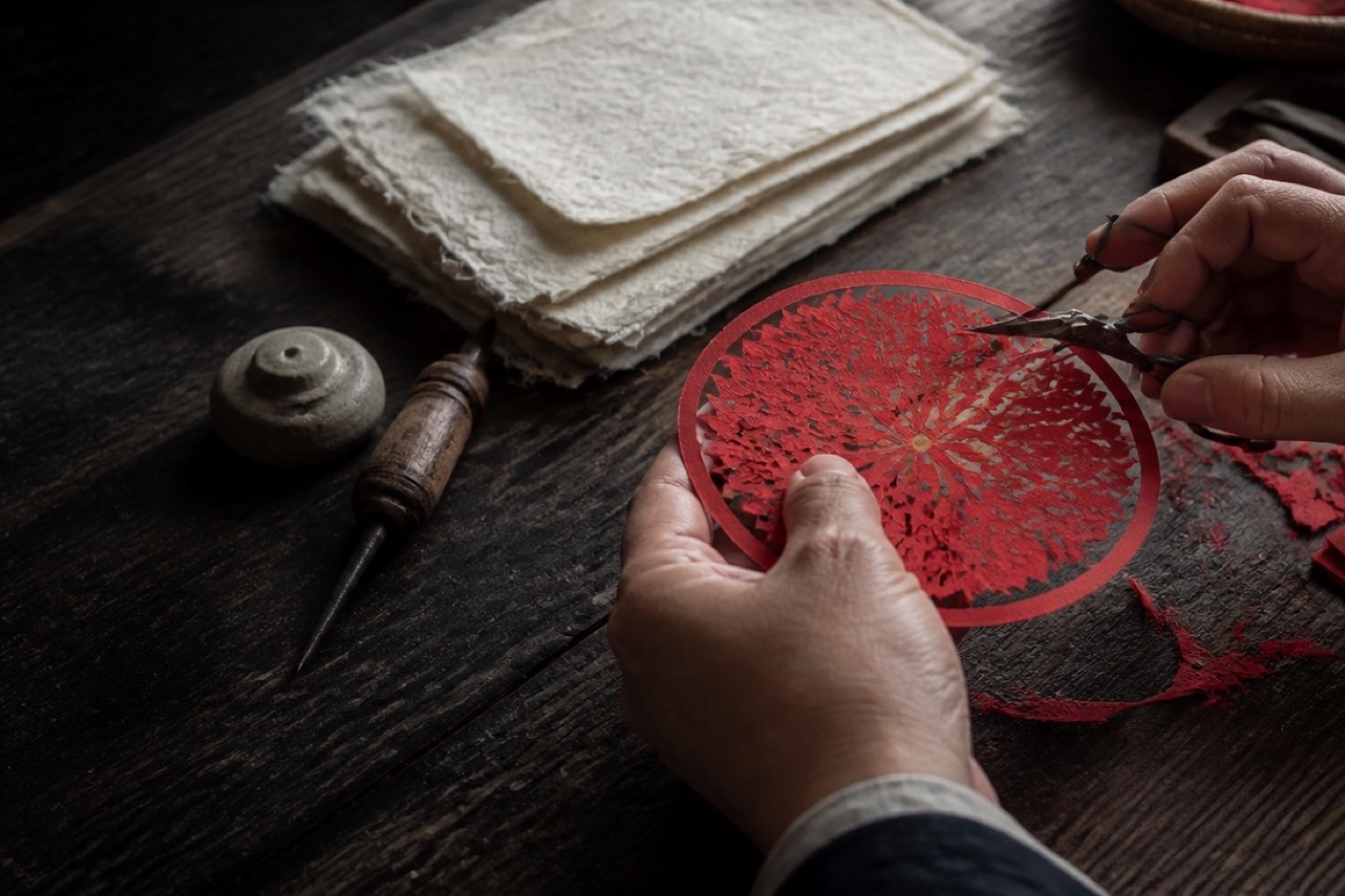 Hands using fine scissors to cut an intricate circular motif from red paper.