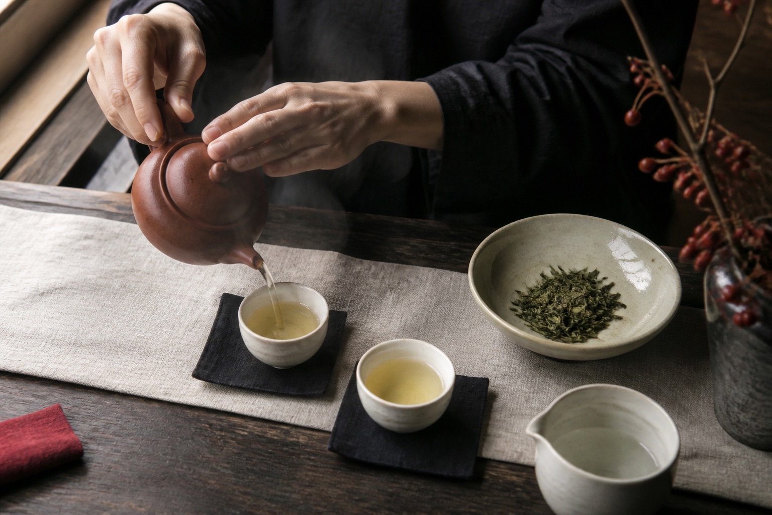 Hands pouring tea from a small clay teapot into simple ceramic cups.