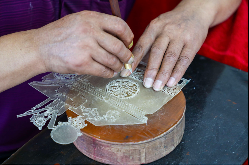 A craftsperson carves intricate patterns into a translucent leather shadow-puppet piece on a wooden block.